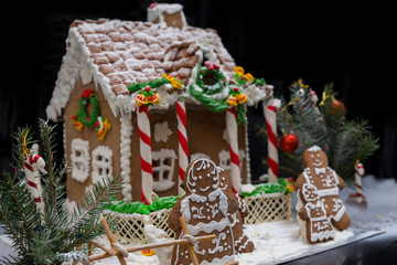 Adorable gingerbread family near snow-covered homemade gingerbread house with sprig of Christmas tree on dark background