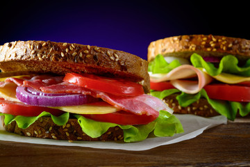 Sandwiches with ham, bacon, tomato, cucumber, cheese, and herbs on dark background.
