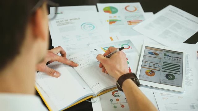closeup businessman writing pen male hand noting copybook paper graph workplace tablet graphs charts digital display pc table commercial working finance app diagram notebook notes employee strategy