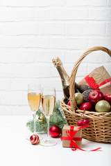 Two glasses of champagne with Christmas basket with bottle, gifts with red satin ribbon, candy canes, pine cones, golden garlands on white brick wall background, new year celebration concept