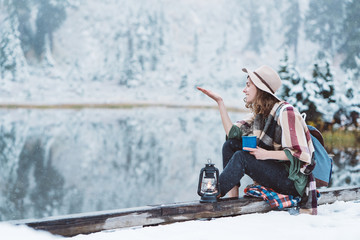 Smiling pretty woman with hot drink catching snow and spending vacations among stunning winter landscape. Traveling in mountains wilderness. Wanderlust and boho style