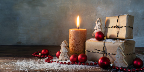 two gift boxes and a burning candle, red christmas baubles and small wooden toy trees in some snow on rustic wood, vintage background with large copy space, panorama format