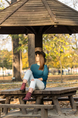 Beautiful teenage girl sitting on the park bench with black hat blue sweater and ripped jeans with blurred autumn leaves in the background