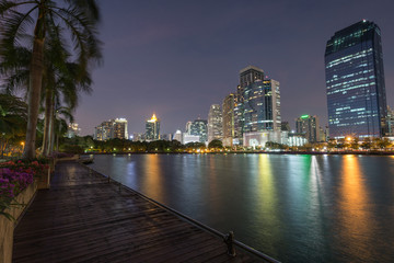 Scenic view of palm trees, wooden boardwalk and lake at the Benjakiti (Benjakitti) Park and lit skyscrapers in Bangkok, Thailand, in the evening. © tuomaslehtinen