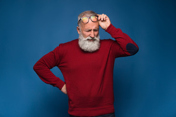 Headache. Old bearded man in red sweeter and in black glasses show headache. Pain emotions isolate on blue background. Stress after hard working office day