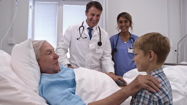 Physician and nurse standing at bedside and looking at happy senior patient holding hand and talking with little grandson