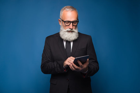 Concentrate bearded senior using internet on a tablet and working in a comfortable way. Serious man in a glasses and modern strict suit