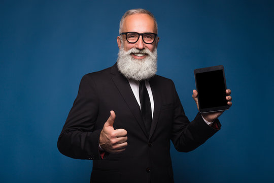 Happy bearded man in formal wear holds digital tablet and shows copy space on screen while standing on blue background. Empty place of screen of tablet. Man show with the screen of tablet and thumb up