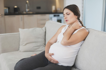 Obraz na płótnie Canvas A pregnant woman sits at home on a light sofa. She has a stiff neck and she rubs it.
