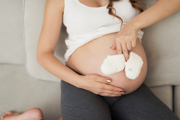 A pregnant woman lies on a light sofa at home. She put baby booties to her belly