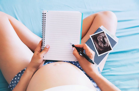 Pregnant woman makes notes in notebook and holding ultrasound image