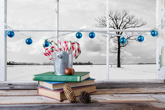 horizontal image of a tin pail sitting on top of books filled with Christmas candy canes in front of window with blue balls strung across the frame with a winter scene on the outside.