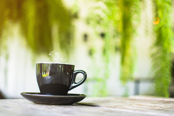 Black cup with steaming hot coffee on the wooden table with blurred green garden background
