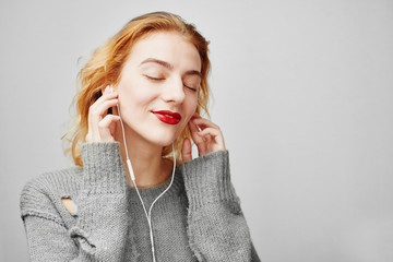 Portrait of a young red-haired girl on a gray background in a sweater, and enjoy listening to music with headphones