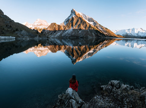 Young adult woman wearing red jacket enjoying the view over a mirror-like mountain lake in the Austrian Alps during sunset