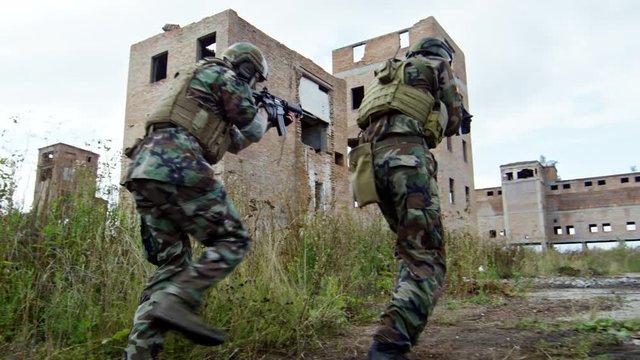 SLOWMO rear view of military team in camouflage running with weapons towards destroyed buildings