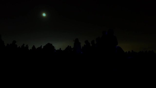 Silhouettes of many people and children holding glowing sticks waiting for fireworks in the dark field, full moon in the sky