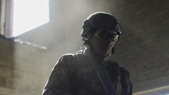 Slowmo of special forces soldier in balaclava and protective glasses walking with rifle in abandoned building; dust floating in sunlight
