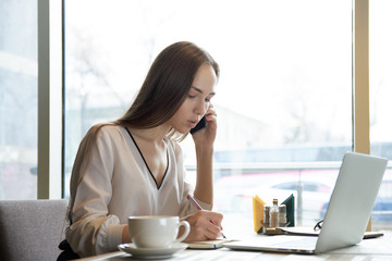 young attractive confident successful woman business woman talking on the phone and making notes in notebook sitting at a table in a cafe with laptop