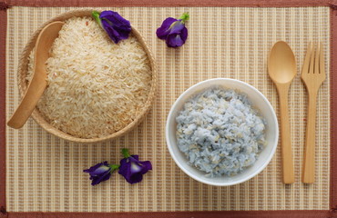 Organic brown raw rice in bamboo basket with wooden spoon and cooked blue rice from butterfly pea flower in white bowl with wooden spoon and fork on bamboo mat,top view