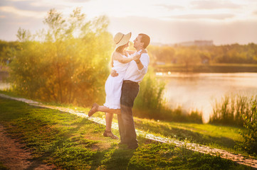 Beautiful bright fairy walk of a loving couple in the summer at sunset. A guy in a white shirt and jeans with a girl in a sarafan and a straw hat hugs and dances against the background of a pond in th