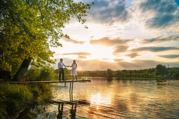 Fototapeta na wymiar Beautiful bright fairy walk of a loving couple in the summer at sunset. A guy in a white shirt and jeans with a girl in a sarafan embraces, dancing, sitting on the pier. Pond in the background. Place