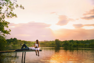  Beautiful bright fairy walk of a loving couple in the summer at sunset. A guy in a white shirt and jeans with a girl in a sarafan embraces, dancing, sitting on the pier. Pond in the background. Place