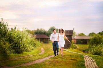 Beautiful bright fairy walk of a loving couple in the summer at sunset. A guy in a white shirt and jeans runs after the girl in a sundress against the backdrop of a watermill in the background. Place 