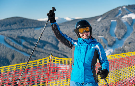 Smiling girl enjoying ski holiday standing on the snowy mountain and raised her hand up at sunny day. Woman at ski resort wearing helmet, blue ski suit and goggles. Blurred background