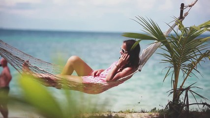 Happy beach woman wearing sunglasses talking mobile phone lying on hammock relaxing on tropical vacation with amazing sea view. 1920x1080