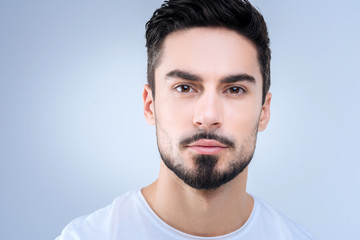 Nice portrait. Young handsome man with a black beard looking calm and concentrated while standing against the blue background