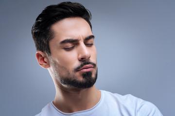 Feeling calm. Relaxed unemotional young man sitting with his eyes closed and meditating while being absolutely alone at home