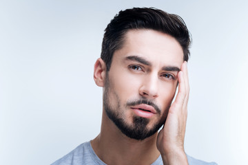 Naklejka premium Unpleasant headache. Ill unhappy young man standing against the blue background and touching his head while having a terrible headache