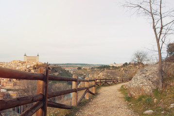 A path with wooden rural fence over the hill with a view to Toledo Alcazar, evening at surroundings of Toledo, Castilla la Mancha, Spain.