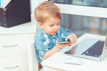 Two years old baby boy talking by phone sitting at desk with laptop near the window in toy office