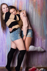Two attractive teenage girls hugging over painted wall. Wearing pullover, jeans shorts and spats....