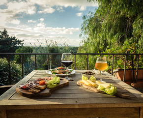 Appetizer for two on a terrace with panorama view.  Beer glass, wine glass, cheese, olives, bread, lettuce and ham on a wooden table