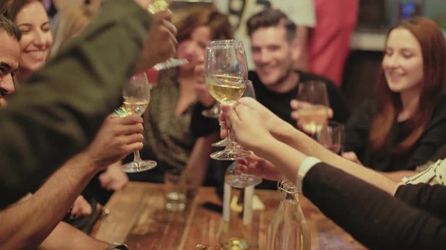 Group of happy, smiling and positive, drunk friends drink and cheer their glasses at party or reunion in middle of crowded popular hipster bar. They laugh and celebrate friendship or new year