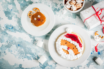 Idea for a children's Christmas breakfast: pancakes decorated like Santa Claus and deer, cocoa with teddy bears and deer marshmallow. On a light blue background, with xmas gift top view copy space