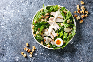 Classic Caesar salad with grilled chicken breast and half of egg in white ceramic plate. Served with croutons and dressing over dark gray texture background. Top view, space.