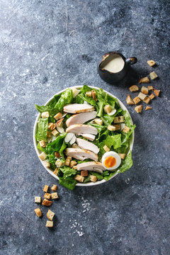 Classic Caesar salad with grilled chicken breast and half of egg in white ceramic plate. Served with croutons and dressing over dark gray texture background. Top view, space.