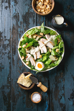 Classic Caesar salad with grilled chicken breast and half of egg in white ceramic plate. Served with ingredients above over old dark blue wooden background. Top view, space.
