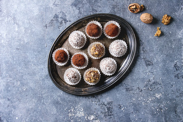 Variety of homemade dark chocolate truffles with cocoa powder, coconut, walnuts on vintage tray over blue texture background. Top view, copy space.