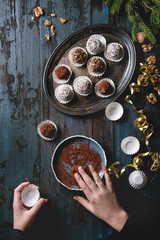 Child hands make homemade dark chocolate truffles with cocoa powder, coconut, walnuts as Christmas gift, put on vintage tray. Fir tree, Christmas decorations above on old wooden table. Top view.
