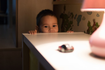 child hiding behind a dresser on which stands a table lamp and toy machine