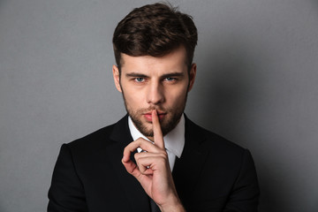 Close-up photo of young handsome man in black suit showing silence gesture, looking at camera