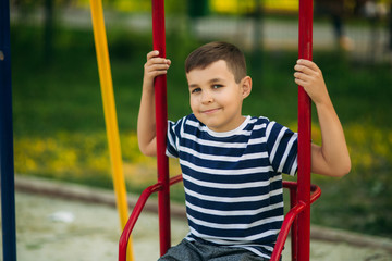 A little boy in a striped T-shirt is playing on the playground, Swing on a swing. Child is smiling and cheering
