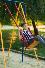 A little boy in a striped T-shirt is playing on the playground, Swing on a swing. Child is smiling and cheering