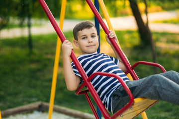 A little boy in a striped T-shirt is playing on the playground, Swing on a swing. Child is smiling and cheering