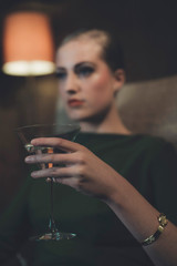Retro 1960s woman sitting with cocktail glass in chair of hotel room at night.